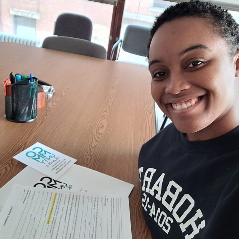 A black woman with short black hair sits at a table with paperwork in front of her. She is smiling at the camera while taking a selfie.