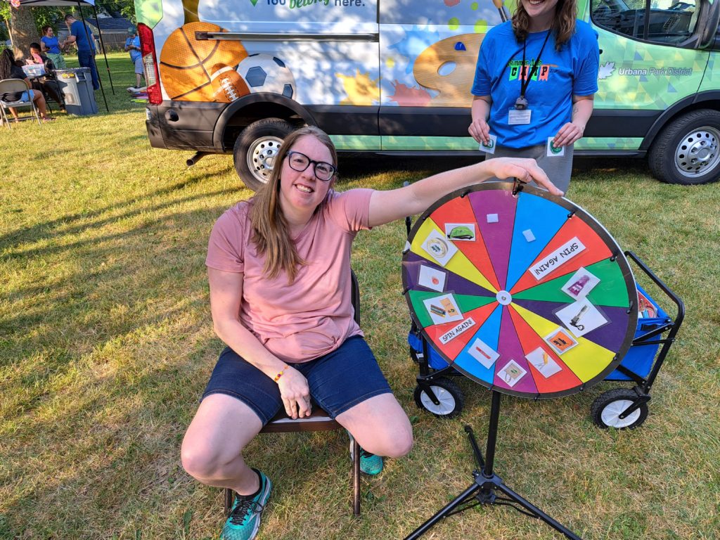 White woman with blonde hair smiles at camera with arm draped over prize wheel at Play Day